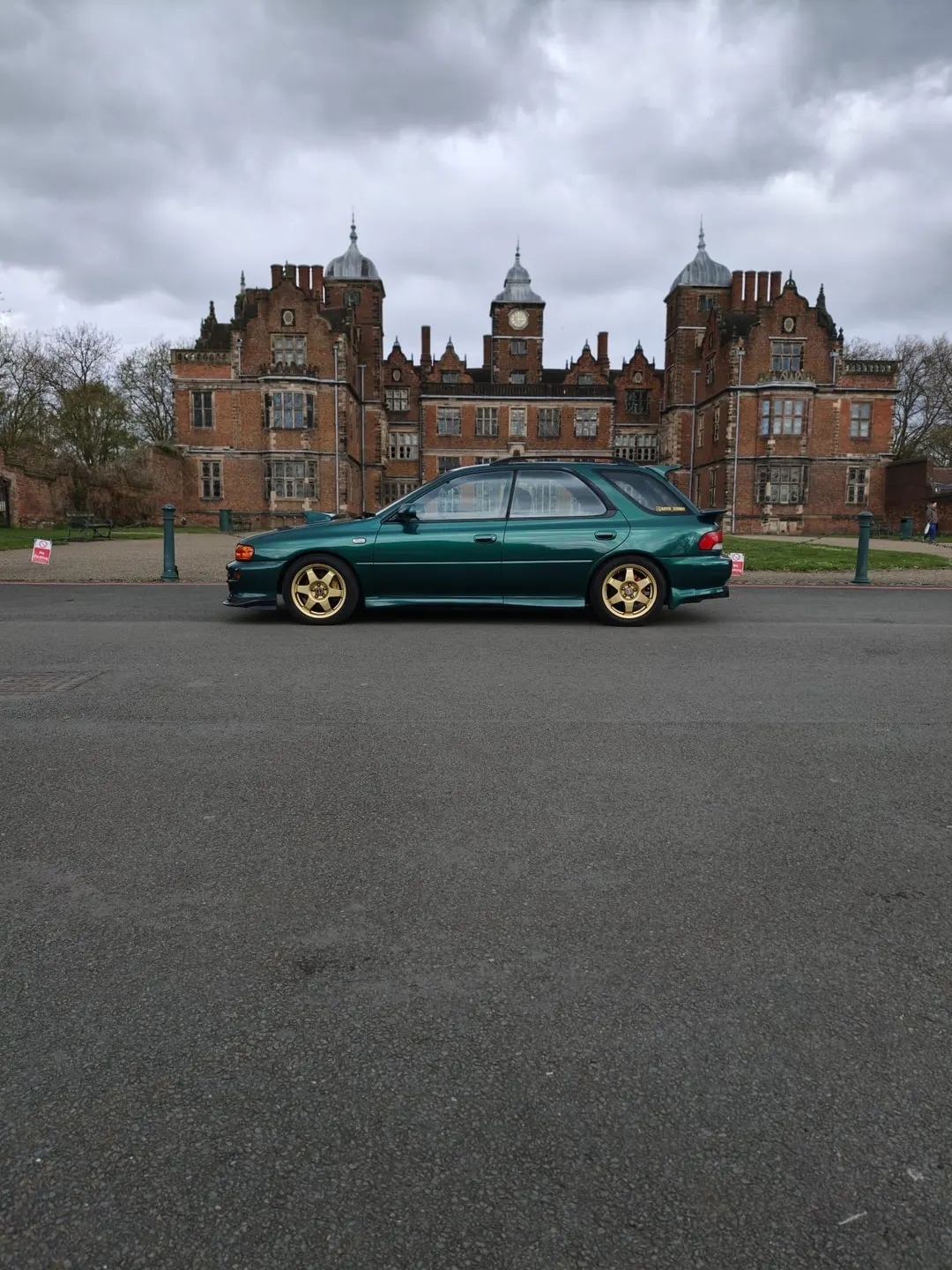 Green Subaru Impreza estate with gold wheels in front of a historic red-brick hall